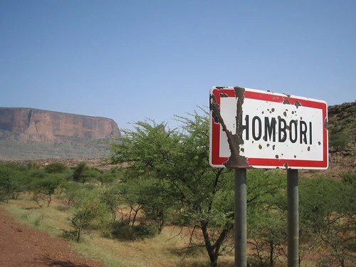洪博里道路旁白底紅框的HOMBORI聚落入口牌，後方可見砂岩崖壁。
