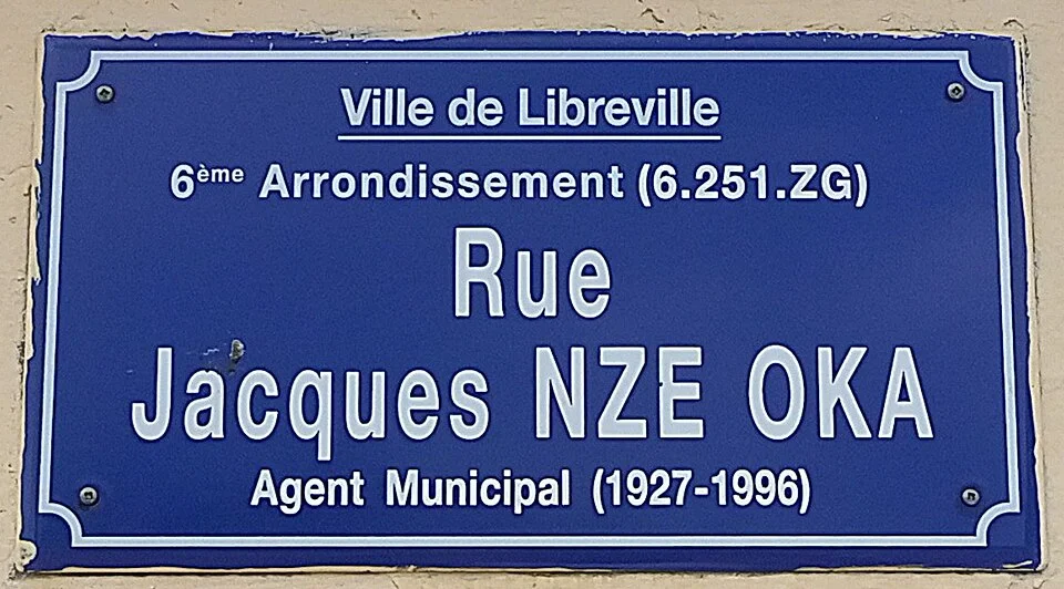 利伯維爾的實拍街牌或門牌。藍牌寫Rue Jacques NZE OKA，上方列Ville de Libreville及6ème Arrondissement (6.251.ZG)，下方標Agent Municipal (1927–1996)。