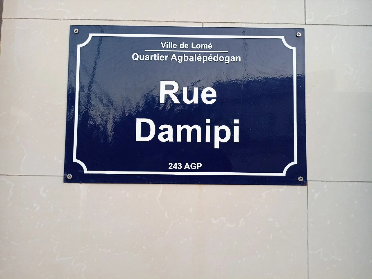 洛梅的實拍街牌或門牌。藍牌主文是Rue Damipi，頂端寫Ville de Lomé及Quartier Agbalépédogan，下方另有243 AGP。