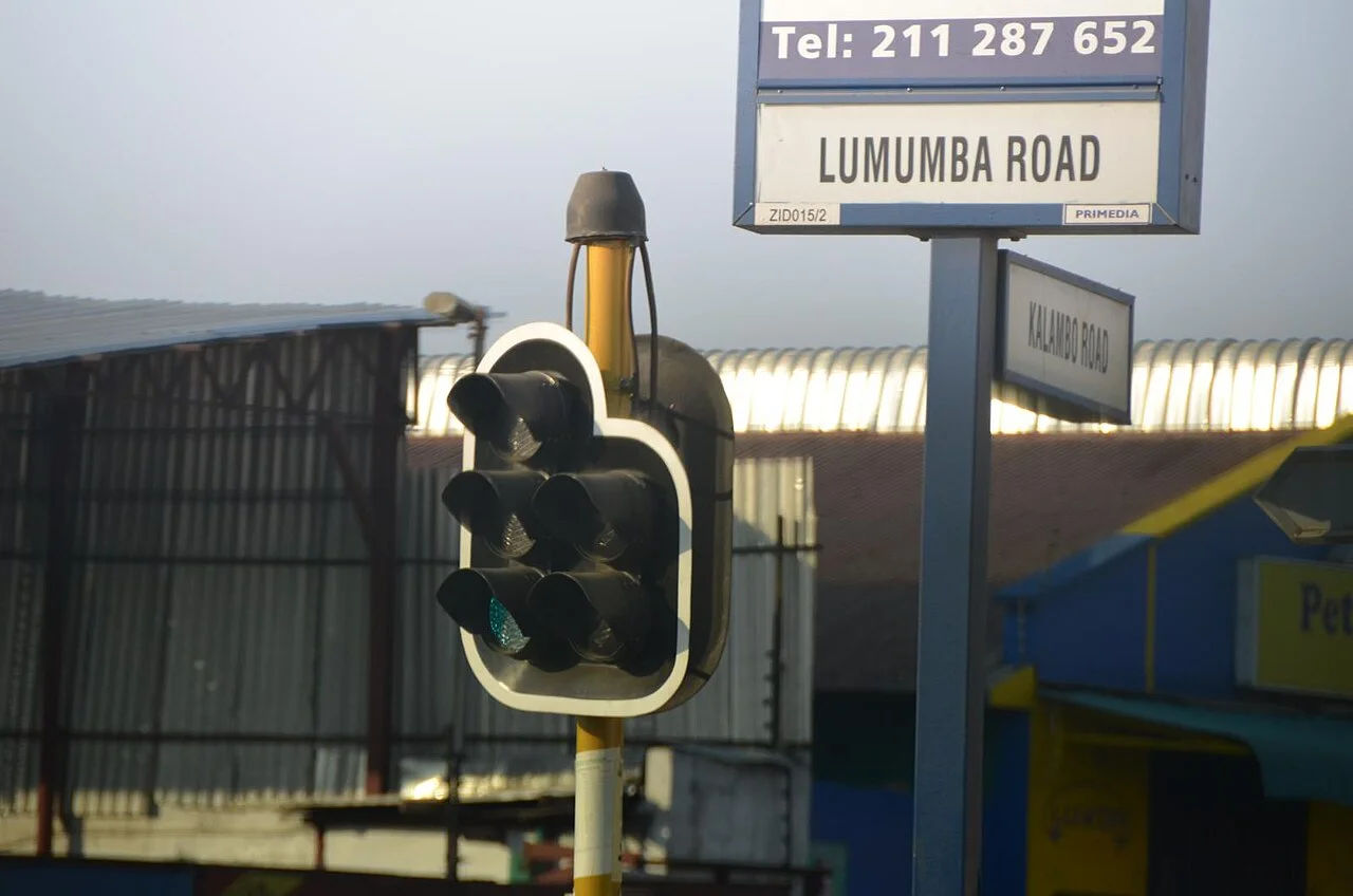 路沙卡的實拍街牌或門牌。交叉路牌分別標LUMUMBA ROAD與KALAMBO ROAD，上方另設電話號碼廣告區。