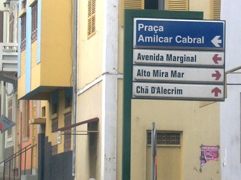 明德盧的實拍街牌或門牌。牌面列Praça Amílcar Cabral及Avenida Marginal，另有Alto Mira Mar與Chã D’Alecrim，各行以箭頭指引方向。