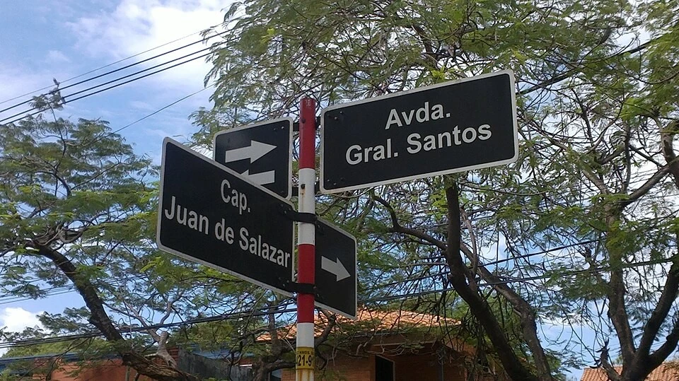 亞松森街名標記。可讀Avda. Gral. Santos與Cap. Juan de Salazar；Avda.、Gral.、Cap.為道路與軍階縮寫，箭頭另表示方向。