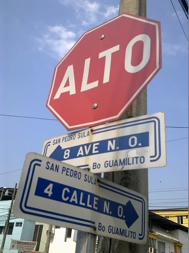 聖佩德羅蘇拉街名標記。可讀8 AVE N.O.、4 CALLE N.O.、SAN PEDRO SULA及Bo GUAMILITO；AVE是Avenida縮寫，N.O.為西北方位縮寫，Bo標示街區。上方ALTO是停止指示。