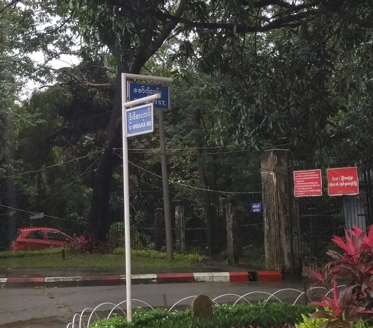 仰光：前牌清楚寫「U WISARA RD」，後牌可辨ZAWGYI ST.但部分被支架遮擋；兩牌上行皆為緬文，未完整轉錄。