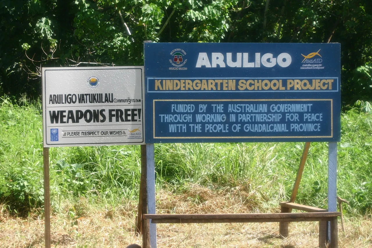 所羅門群島阿魯利戈路旁並列ARULIGO-VATUKILAU社區和平公告白牌與ARULIGO幼兒園援建藍牌。