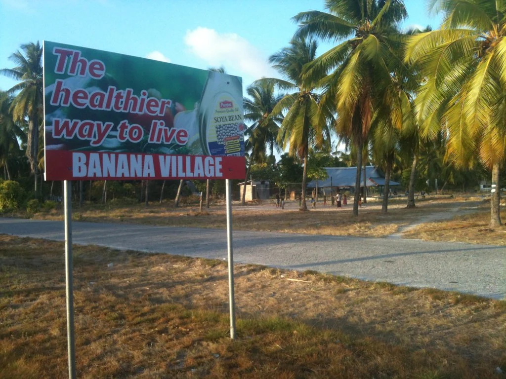 吉里巴斯巴納納村路旁看板，下沿紅底白字BANANA VILLAGE，背景為道路、椰樹與民居。