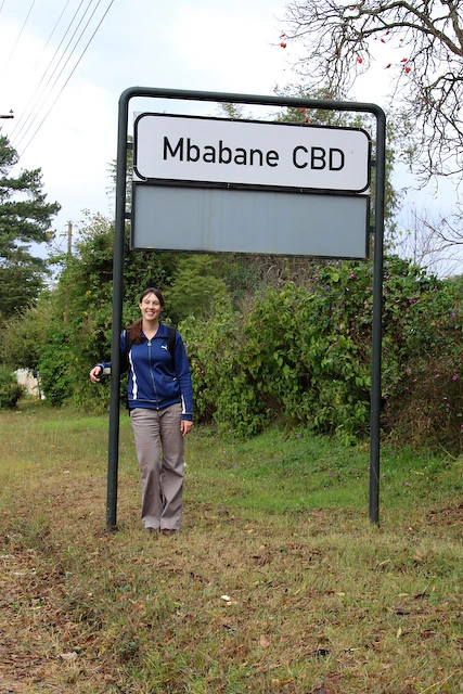 白底黑框的Mbabane CBD商業中心區地方牌，雙立柱旁有行人，背景為草地、樹木與電線。