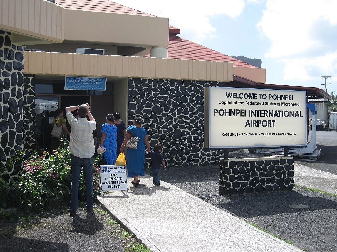 波納佩航站入口白牌頂部寫WELCOME TO POHNPEI，下方列機場名稱和四種問候語，旁邊有抵達旅客與石砌牆面。