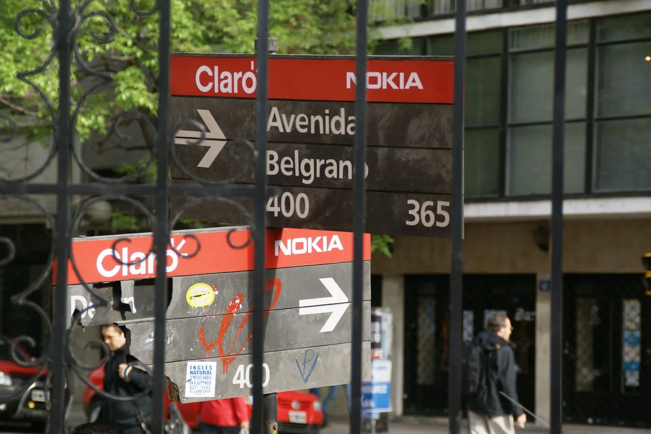 從布宜諾斯艾利斯修道院鐵欄後望見黑底白字Avenida Belgrano街牌、右向箭頭及周圍紅色廣告。