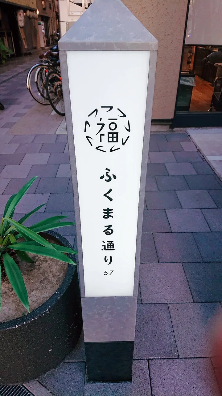 大阪福島的ふくまる通り57直立街道標示，白色面板上有圓形福字標誌、直排街名及57。