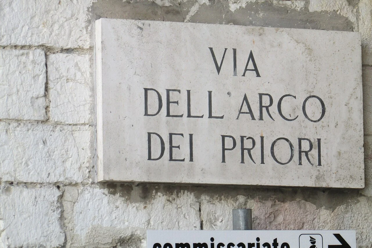 阿西西的實拍街牌。「VIA DELL’ARCO DEI PRIORI」為完整街名,分成四行;下方其他指示另屬畫面資訊。