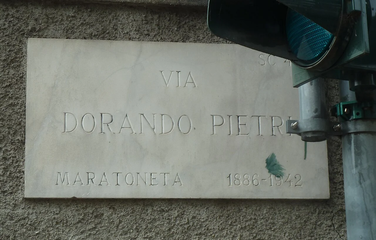貝加莫的實拍街牌。「VIA DORANDO PIETRI」為街名；「MARATONETA 1886–1942」是馬拉松選手的職業與生卒年。
