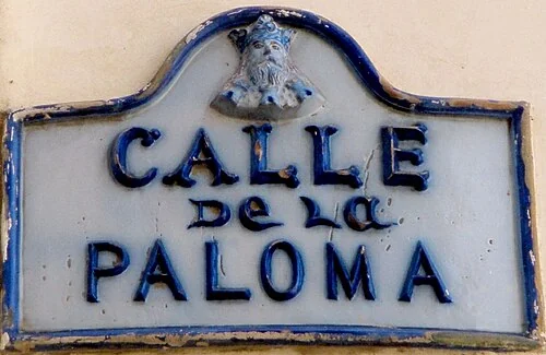 布哥斯的實拍街牌。「CALLE DE LA PALOMA」為街名；上緣鴿子形象與Paloma的字義相呼應，不能單憑圖像確認語源。