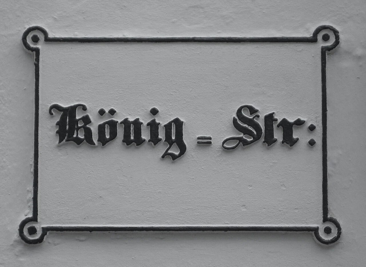 呂貝克的實拍街牌。牌面「König-Str:」以縮寫表示Königstraße；冒號狀標點與黑體字是牌面可見特徵。