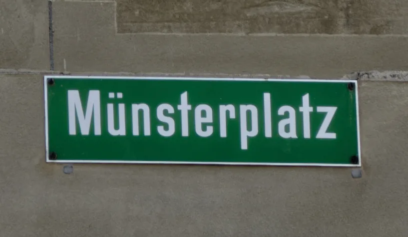 伯恩石牆上的綠色Münsterplatz路牌，白字白框，四角以深色螺絲固定。