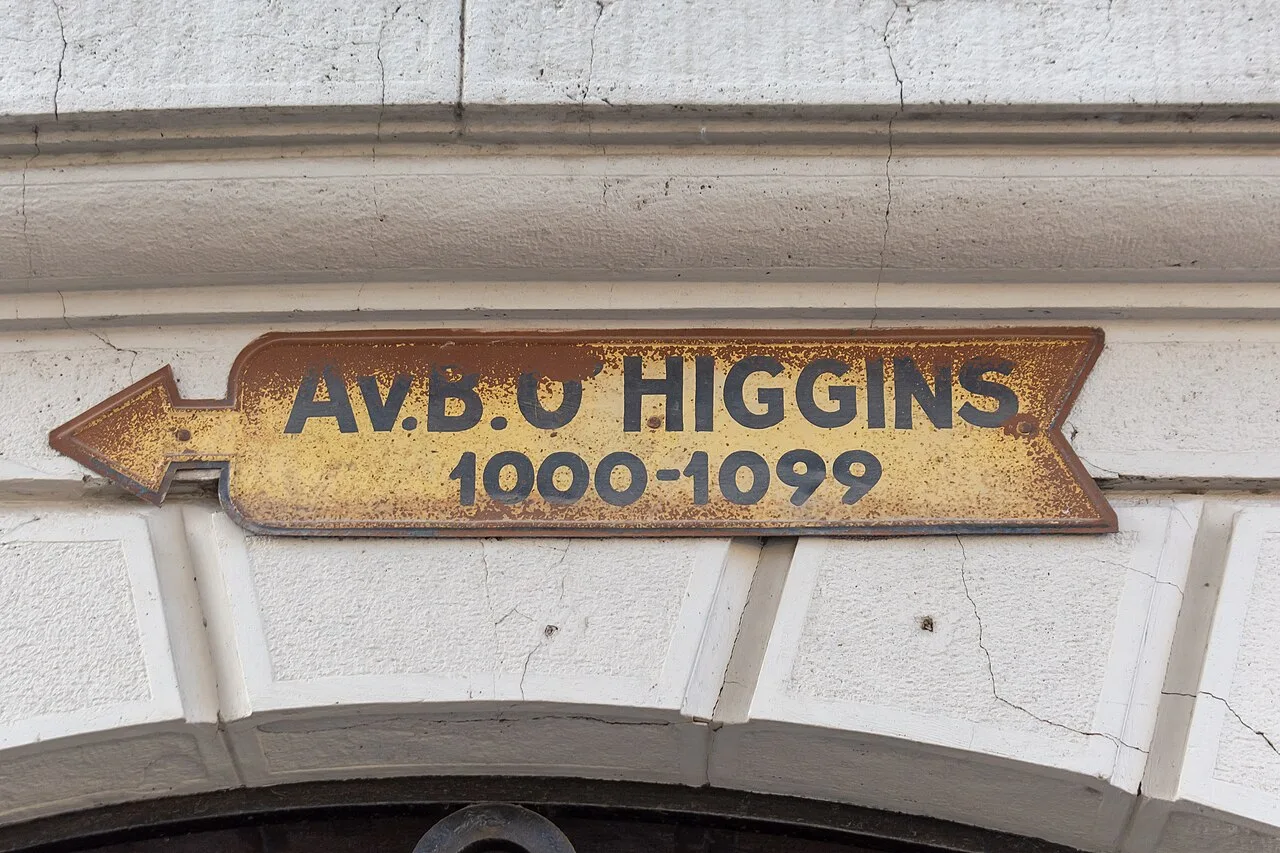 聖地牙哥建築外牆上的黃色箭頭街牌,寫Av. B. O’HIGGINS與1000–1099,表面有鏽色磨損。