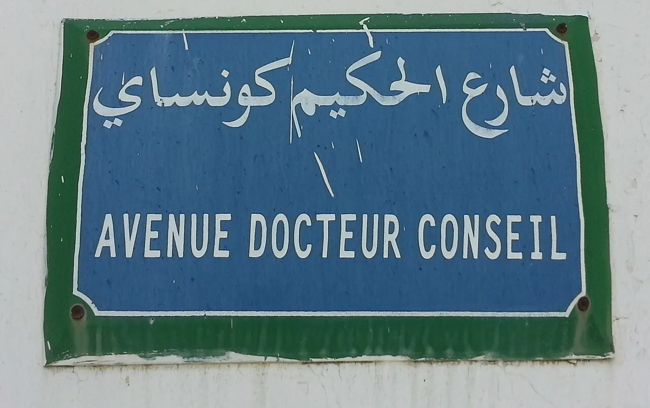 突尼斯市藍底白字街牌,上排阿拉伯文,下排AVENUE DOCTEUR CONSEIL,外圍為綠框與四角螺釘。