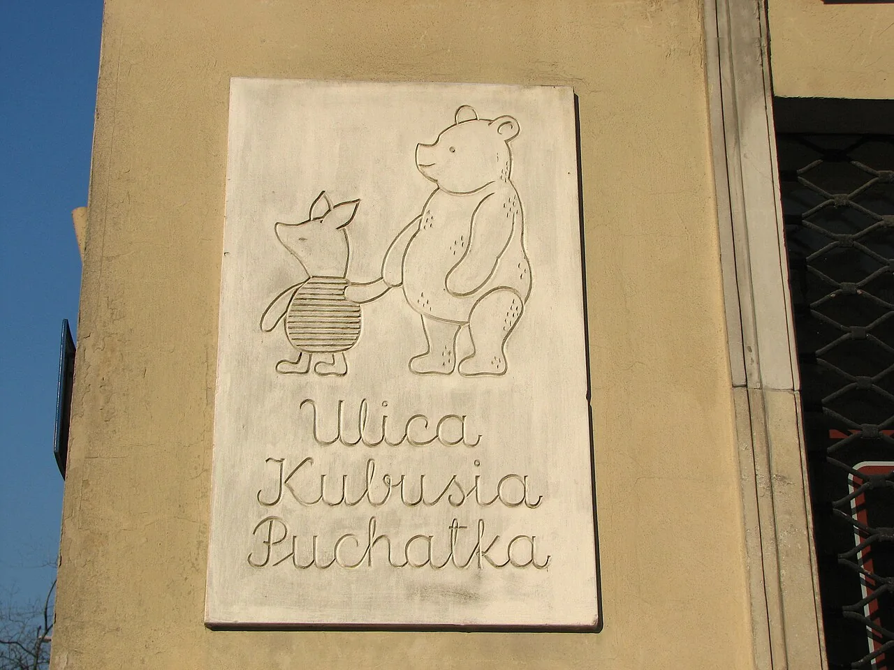 華沙小熊維尼街的米色浮雕牌，小熊維尼與小豬位於Ulica Kubusia Puchatka字樣上方。