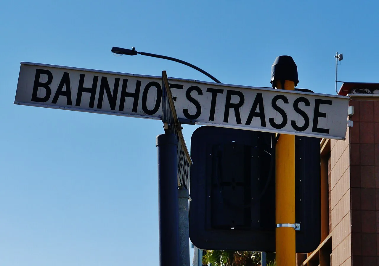 溫荷克白底黑字BAHNHOFSTRASSE長牌，前方有交叉街牌側面，背景為藍天與號誌背板。