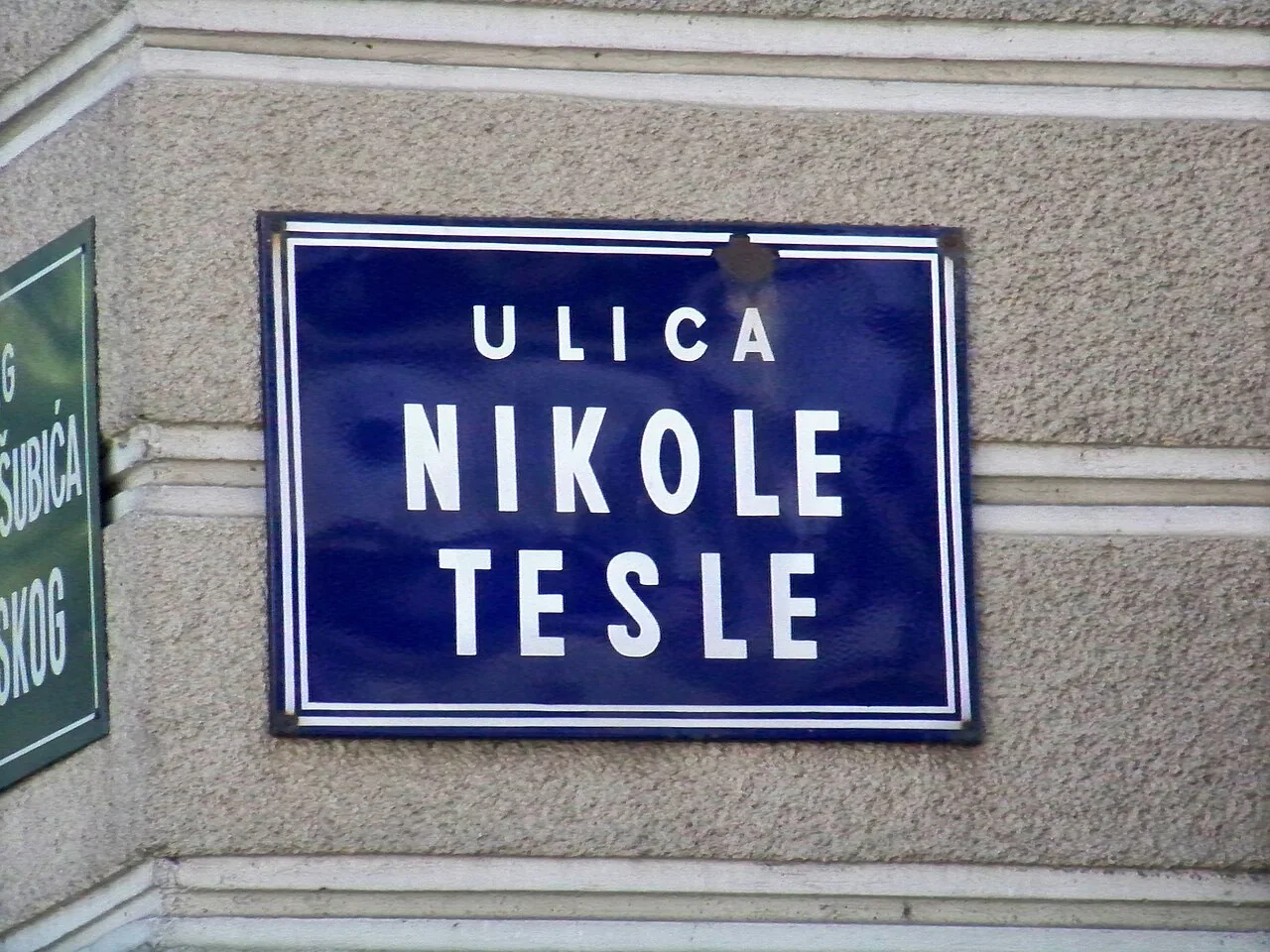 札格雷布灰牆上的藍色雙白框路牌，白色大字分行寫ULICA NIKOLE TESLE。