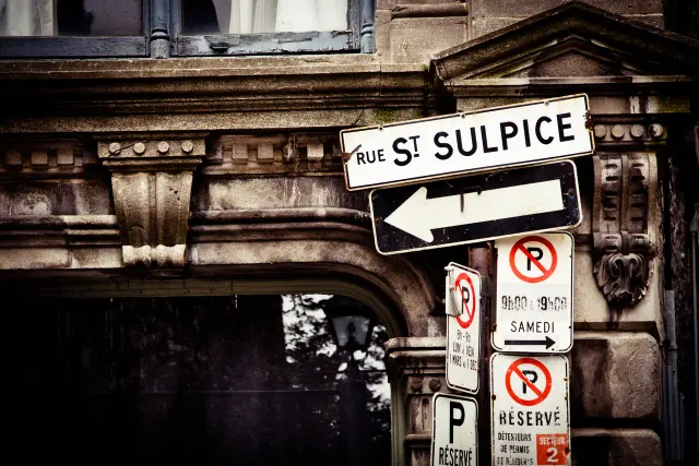蒙特婁白底黑字Rue St Sulpice街牌，底下有向左單行箭頭及停車規則牌，後方是石造建築。