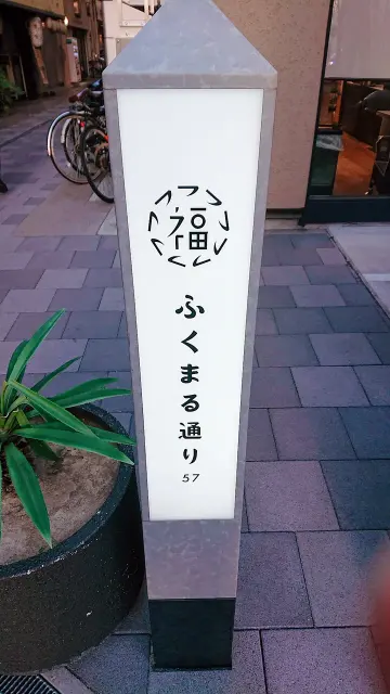 大阪福島的ふくまる通り57直立街道標示，白色面板上有圓形福字標誌、直排街名及57。