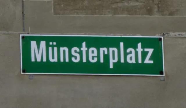 伯恩石牆上的綠色Münsterplatz路牌，白字白框，四角以深色螺絲固定。