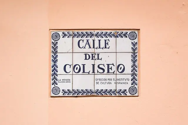 波哥大粉橘色牆上藍白拼接磚牌，三行寫CALLE DEL COLISEO，外圍有花葉紋，底部有捐贈說明。