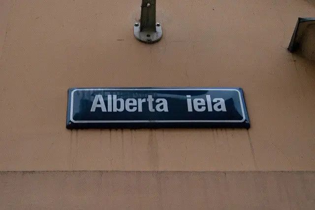 里加赭黃牆上的深藍Alberta iela路牌，白色粗字與圓角白色邊框。