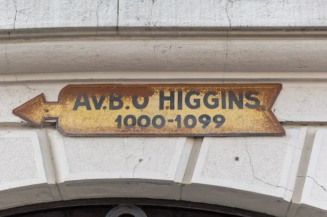 聖地牙哥建築外牆上的黃色箭頭街牌，寫Av. B. O’HIGGINS與1000–1099，表面有鏽色磨損。
