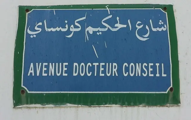 突尼斯市藍底白字街牌，上排阿拉伯文，下排AVENUE DOCTEUR CONSEIL，外圍為綠框與四角螺釘。
