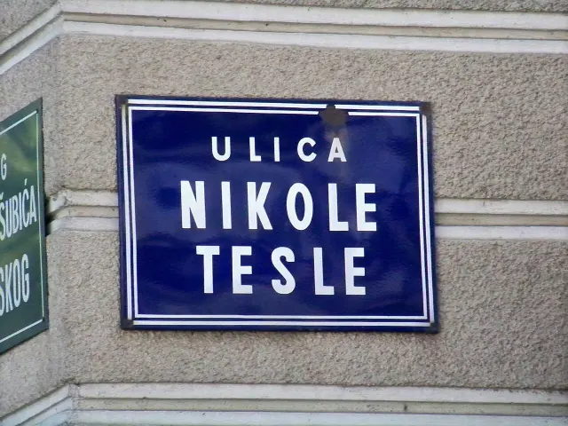 札格雷布灰牆上的藍色雙白框路牌，白色大字分行寫ULICA NIKOLE TESLE。