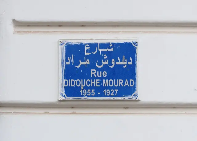 阿爾及爾的實拍街牌。「Rue DIDOUCHE MOURAD」與上方阿拉伯文為街名；下方牌面由左至右寫「1955 - 1927」，對應人物卒年與生年，並非門牌。