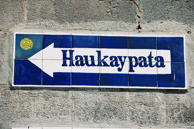 庫斯科藍白Haukaypata指示牌，左側金色圓臉圖案，白箭頭朝左。