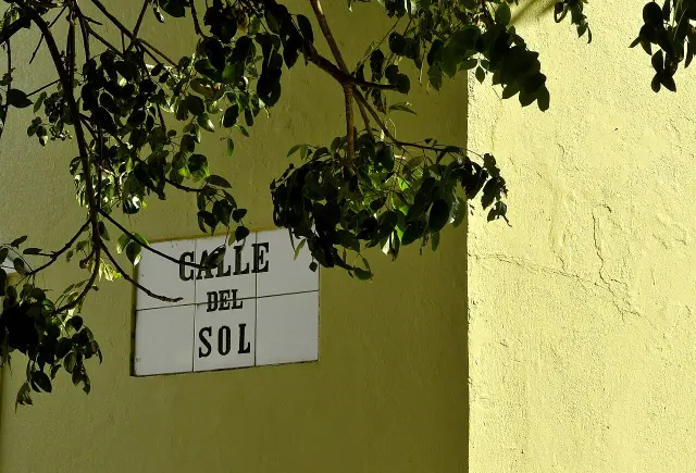 舊聖胡安牆上白磚黑字CALLE DEL SOL街牌，部分表面有樹葉陰影。
