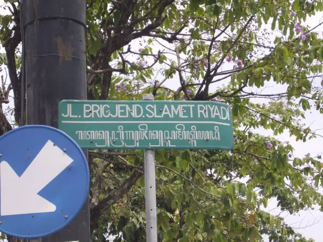 梭羅綠色街牌上排為JL. BRIGJEND. SLAMET RIYADI，下排為爪哇文字，前方部分被藍色交通箭頭牌遮住。
