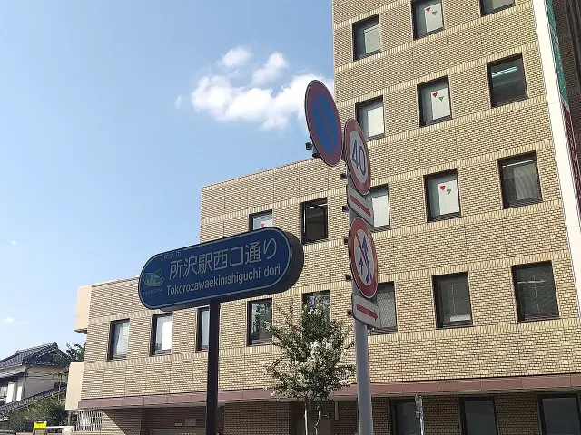 所澤站西口通的藍色日文及羅馬字街牌，後方是淺色磚面建築。