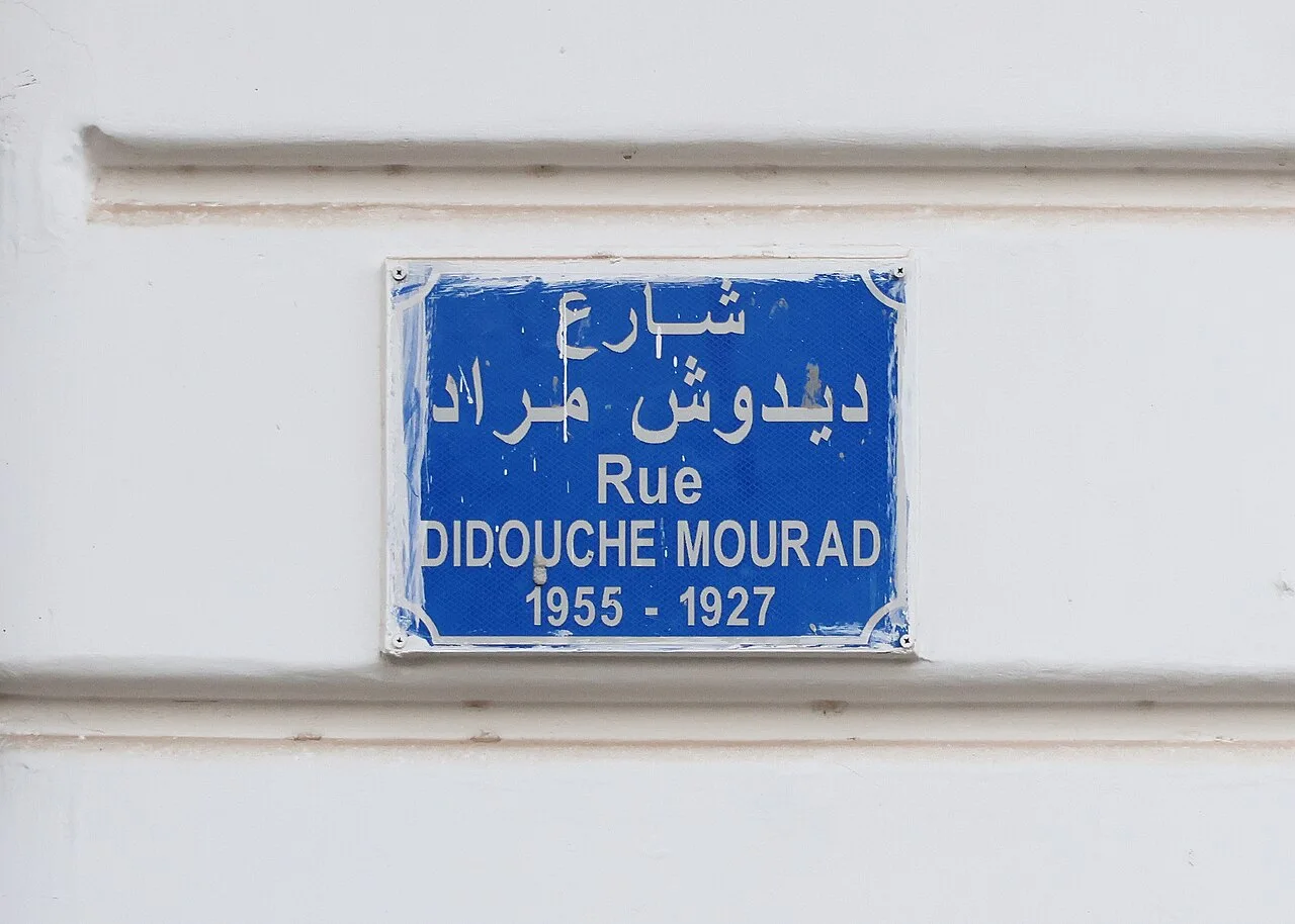 阿爾及爾的實拍街牌。「Rue DIDOUCHE MOURAD」與上方阿拉伯文為街名;下方牌面由左至右寫「1955 - 1927」,對應人物卒年與生年,並非門牌。