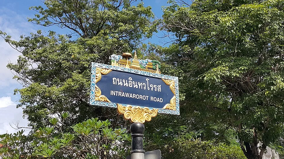 清邁樹下的因他瓦羅羅路牌,泰文與INTRAWAROROT ROAD白字配藍底,外有金色捲紋和塔形飾件。