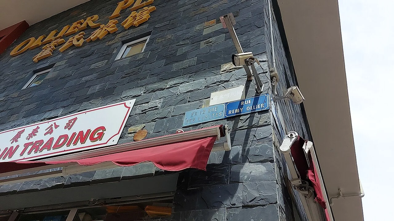 路易港石牆上的藍色RUE REMY OLLIER街牌,旁邊保留EX 12 RUE DES LIMITES舊牌,上方有淡色漢字。