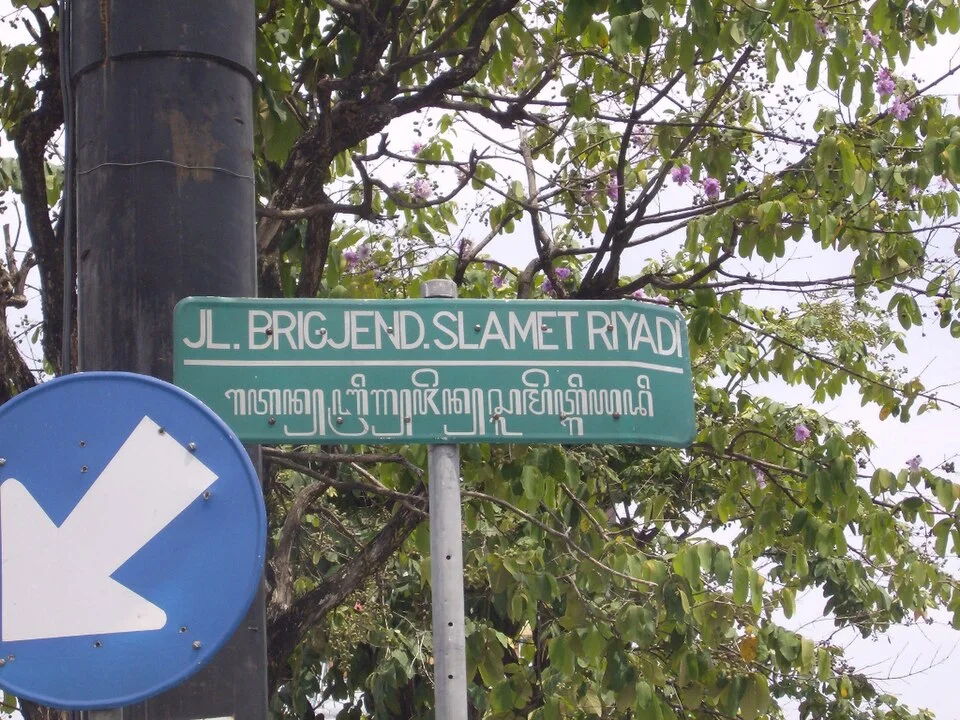 梭羅綠色街牌上排為JL. BRIGJEND. SLAMET RIYADI，下排為爪哇文字，前方部分被藍色交通箭頭牌遮住。