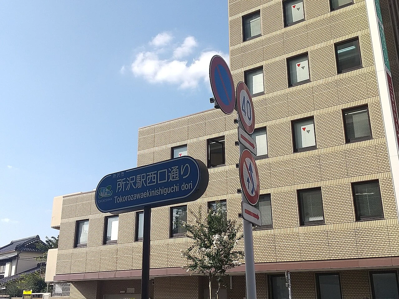 所澤站西口通的藍色日文及羅馬字街牌,後方是淺色磚面建築。